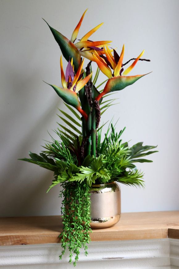 Niezwykła kompozycja z kwiatów strelicji otoczonych egzotycznymi roślinami, ułożona w złotym naczyniu ze szkła.